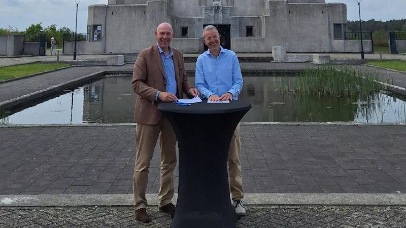 Johan Maas en Mark Vermeer staan glimlachend achter een statafel voor het gebouw van Radio Kootwijk, na het tekenen van de intentieverklaring MijnOverheid Zakelijk.